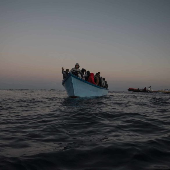 50 Menschen auf blauen Holzboot in Seenot. Im Hintergrund ist die Crew der Sea-Watch 5 mit Rettungsausrüstung zu sehen.