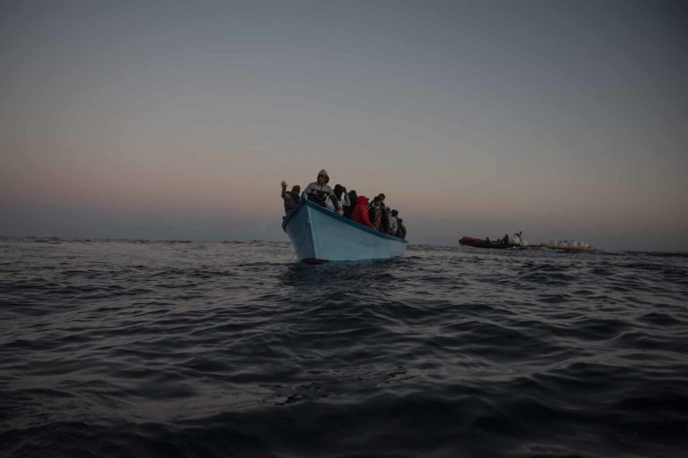 50 Menschen auf blauen Holzboot in Seenot. Im Hintergrund ist die Crew der Sea-Watch 5 mit Rettungsausrüstung zu sehen.